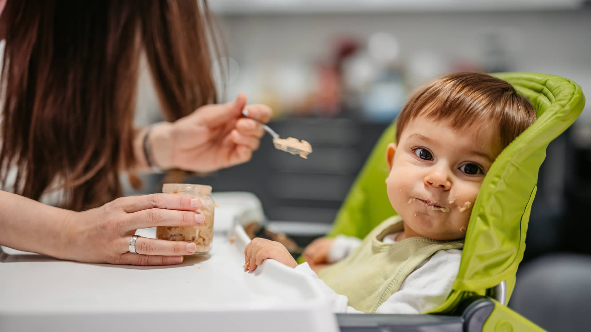Niño pequeño mirando a cámara mientras su madre le da de comer un potito casero con diicultad, hablaremos de la alimentación infantil apropiada para cada edad