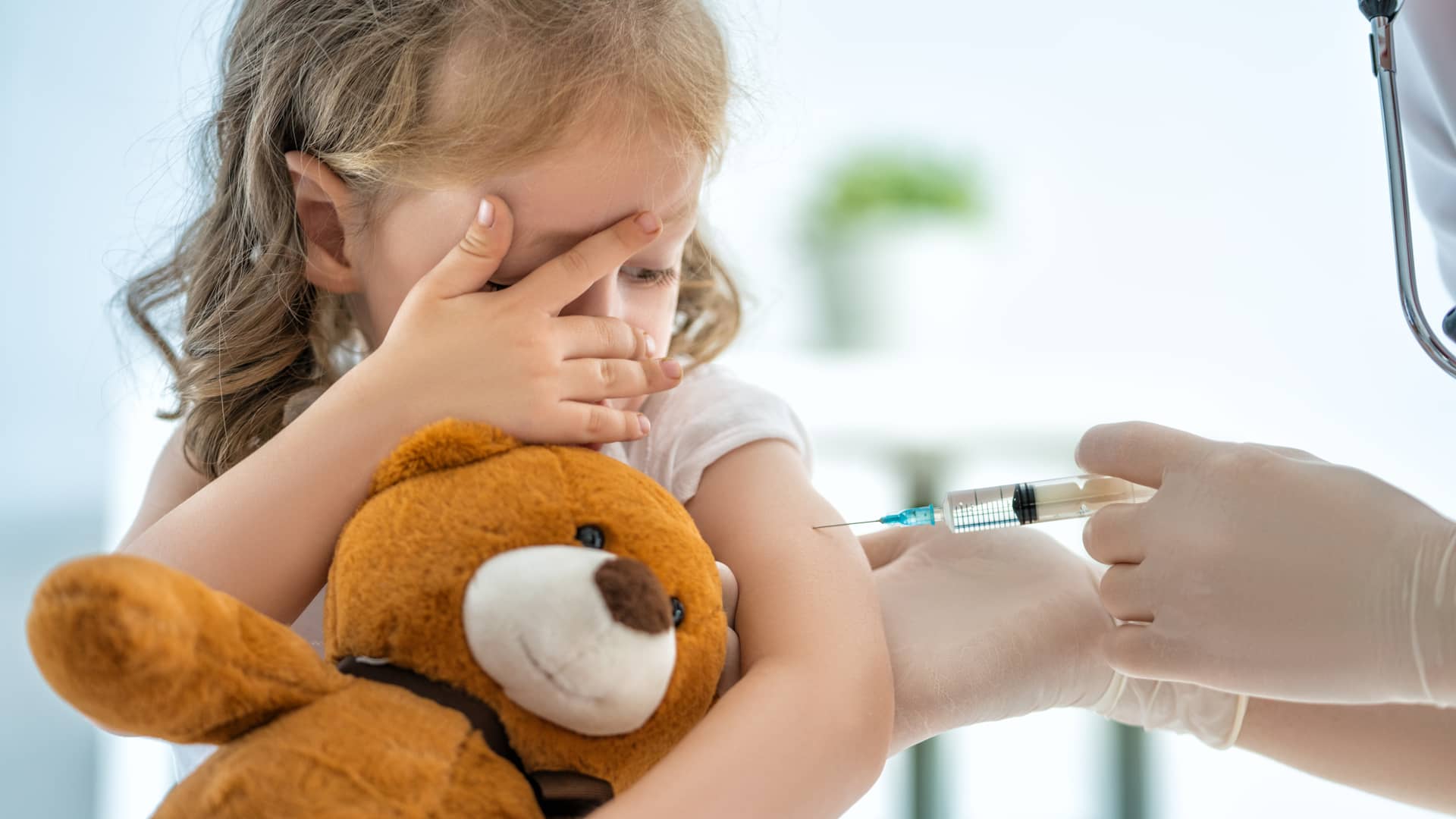 Niña pequeña se tapa la cara y abraza a su oso mientras recibe una vacuna de su pediatra